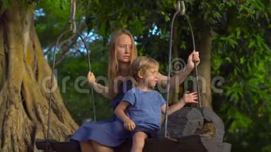 母亲和儿子在热带公园的一棵大热带树上悬挂着一个异国情调的老秋千。 旅行与儿童概念